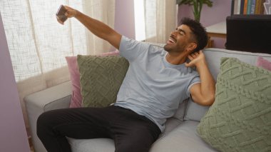 Genç İspanyol bir adam oturma odasında kanepede uzanıp akıllı telefonuyla selfie çekiyor.