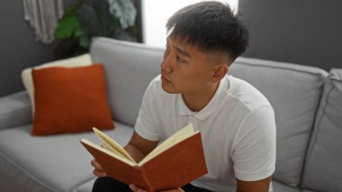 Yakışıklı Asyalı bir adam, gri kanepesi ve turuncu yastığı olan rahat bir oturma odasında oturmuş kitap okuyor.