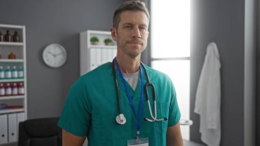 Klinikteki kendine güvenen erkek doktor yeşil önlük ve raflı steteskop takıyor ve arka planda profesyonel bir sağlık hizmeti sunuyor.