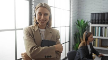 Modern ofis ortamında gülümseyen ve arka planda meslektaşı olan bir kadın, iş ortamında takım çalışması ve iletişimi resmediyor.