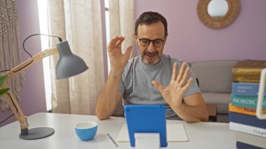Orta yaşlı İspanyol bir adam, rahat bir oturma odasında, elinde bir bardak ve lambayla bir masada otururken el kol hareketleriyle bir video görüşmesi yapıyor..