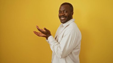 Yakışıklı adam izole edilmiş sarı arka planda gülümsüyor açık gömlekli, açık avuçlu neşeli bir ifade sergiliyor.