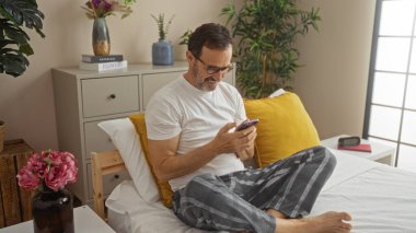 Orta yaşlı, pijamalı, akıllı telefon kullanan, kapalı bir yatak odasında, içinde bitkiler ve dekor olan bir yatakta oturan İspanyol bir adam.