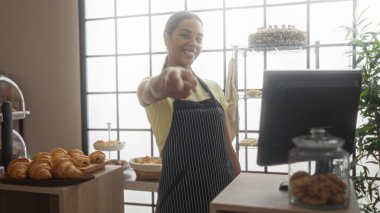 Pastanede önlük takan gülümseyen bir kadın. Kapalı alanda pasta ve pastalar sergileniyor.