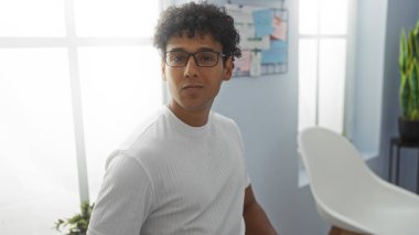 Büyük pencereli, beyaz sandalyeli ve arka planda görünen bitkiler olan parlak bir ofiste oturan İspanyol gözlüklü adam..