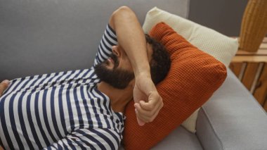 Oturma odasındaki kanepede sakallı genç bir adam. Kolu alnını kapatmış, çizgili bir gömlek giyiyor ve turuncu ve beyaz yastıkların üzerinde yatıyor..