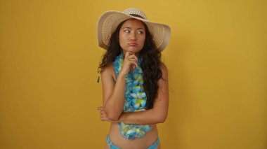 Young woman in blue flower bikini and sunhat stands thoughtfully against a yellow background clipart