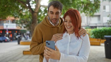 Şehir parkında ağaçlarla çevrili bir kadın ve erkek telefonda gülümsüyor, açık havada neşeli bir anı yakalıyorlar, sevgi ve mutluluk sergiliyorlar..