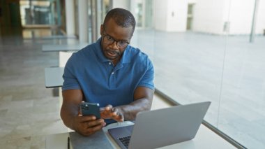 Ofisteki yakışıklı Afrikalı genç adam bilgisayarla çalışırken akıllı telefon kullanıyor, ev ortamında odaklanma ve verimlilik sağlıyor.