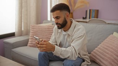 Oturma odasında sakallı bir İspanyol genç akıllı telefon kullanarak rahat bir kanepede, rahat bir evde, pembe yastıklı bir şekilde oturuyor.