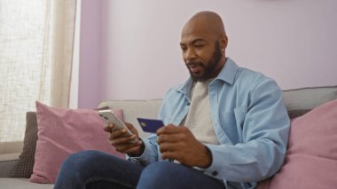 Oturma odasında elinde telefon ve kredi kartıyla oturan sakallı genç adam rahat bir ev ortamında online alışveriş yapmaya odaklanıyor.