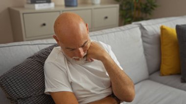 Beyaz saçlı, yakışıklı, kel bir adam yumuşak yastıklar ve sıcak dekorlarla dolu modern bir oturma odasında kanepede kestiriyor. Huzurlu ve rahat bir ev atmosferi sunuyor..