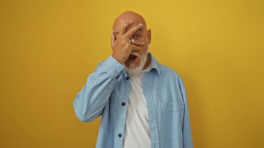 Gri saçlı, sakallı, olgun kel bir adam yüzünü eliyle örterken izole edilmiş sarı bir arka plana şaşkın şaşkın bakıyor..