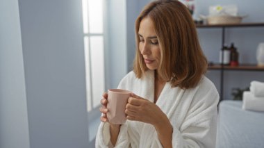 Beyaz bir bornoz giymiş, elinde fincanla kaplıcada dinlenen bir kadın, sağlıklı bir çevreye işaret eden sakin bir iç mekan ile çevrili.