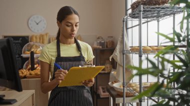 Fırında çalışan genç bir kadın, kafenin iç kısmındaki panoya notlar yazıyor. Pasta ve ekmek raflarıyla.