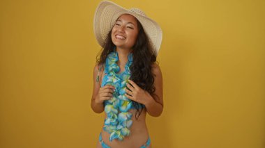 Sarı arka planda duran, şapkalı ve çiçekli, bikinili, gülümseyen genç kadın.