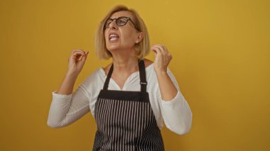 Middle-aged blonde woman in an apron angrily gesturing, isolated against a yellow background, expressing frustration or anger, with short hair and glasses. clipart