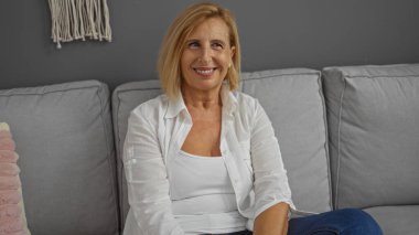 Orta yaşlı, sarışın, beyaz, kısa saçlı bir kadın. Oturma odasında otururken gülümsüyor..