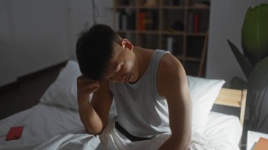 Yatak odasındaki genç adam, elleri başının üstünde, gri atlet giyiyor, ışıkla aydınlatılmış, aşağı bakıyor, ev dekoruyla çevrili, ev dekoruyla çevrili, düşünceli bir şekilde taşınıyor.