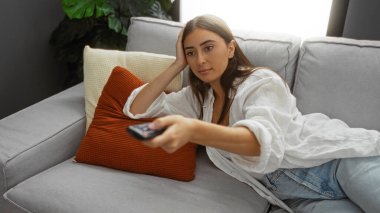 İspanyol kadın oturma odasında uzanıyor, günlük kıyafetler giyiyor, elinde uzaktan kumandayla rahat bir apartman dairesinde minderli bir koltukta dinleniyor..