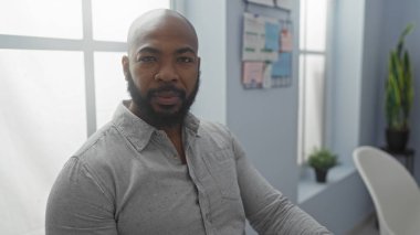 Ofiste oturan sakallı yakışıklı adam duvarında posterler ve penceresinde yeşil bitkiler var.