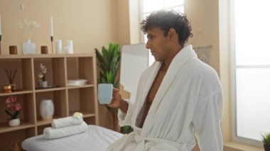 Bir spa ortamında, bornoz giymiş, elinde bir bardak tutan, güzellik ürünleri ve sakin dekorun arasında, sağIıklı bir ortam içinde rahatlayan genç bir İspanyol.