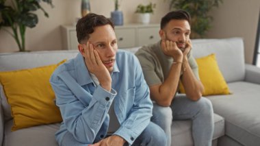 Erkekler birlikte kanepede otururlar. Oturma odasında sıkılırlar. Evde sarı yastıklar vardır..