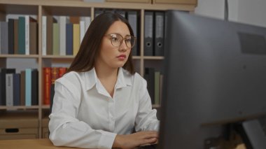 Bilgisayarda bir ofiste çalışan güzel İspanyol bir kadın, arka planda raflar olan modern kapalı bir işyerindeki görevlere odaklanmış..