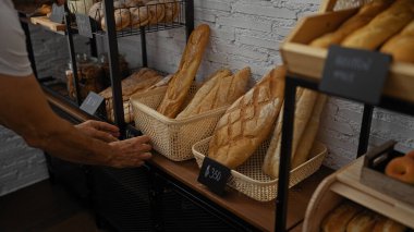 Bir pastanede taze ekmek düzenleyen bir adam. Kapalı alandaki raflarda çeşitli somunlar var. Dolar olarak fiyatlandırılmış..