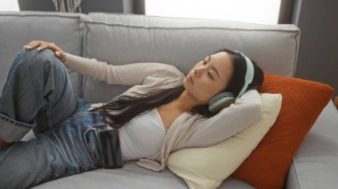 Genç Çinli bir kadın, evindeki oturma odasında, kulaklıklarıyla koltukta dinleniyor, çekici ve huzurlu bir an yaşıyor..