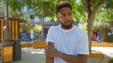 Beyaz tişörtlü genç adam arka planda ağaçlar ve binalar olan güneşli bir şehir parkında dikilirken kolunu kaşıyordu..