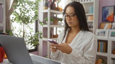 Genç Asyalı bir kadın güzel dekore edilmiş bir ev içi mağazada akıllı telefon kullanıyor, dizüstü bilgisayar üzerinde çalışırken etrafını bitkiler ve dekorasyon malzemeleriyle donatmış.