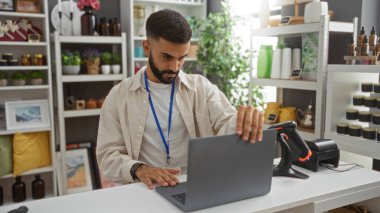 Modern dekorasyon ve aksesuarlarla dolu bir mağazada dizüstü bilgisayarda çalışan yakışıklı İspanyol bir adam.