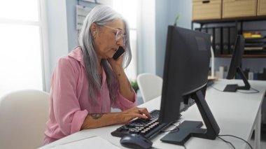 Modern ofis ortamında bilgisayar başında çalışan ve telefonla konuşan bir kadın, iş ortamında çoklu görev ve profesyonel uzmanlık gösteriyor