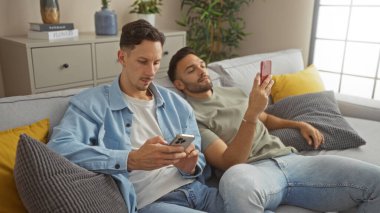 Çift oturma odasındaki koltukta dinleniyor, her biri akıllı telefon kullanıyor, modern teknolojiyle evlerinde rahat ve samimi bir anı tasvir ediyorlar.