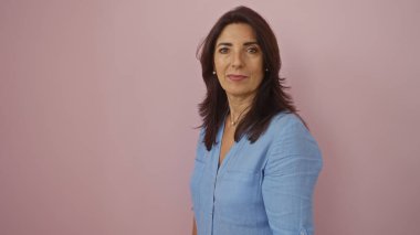 Soyutlanmış pembe arka planda duran, ciddi bir ifadeyle mavi bluz giyen bir kadın.