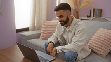 Samimi oturma odasında dizüstü bilgisayara gülümseyen sakallı genç İspanyol adam rahat bir ev ortamında kanepede oturuyor.