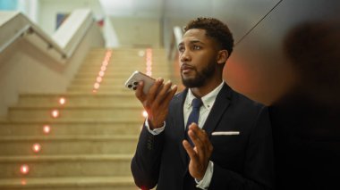Genç iş adamı akıllı telefondan konuşuyor, merdivenlerin yanında duruyor, takım elbise ve kravat takıyor.