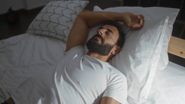 Yakışıklı, sakallı, rahat bir yatak odasında dinlenen, beyaz gömlek giyen ve evde rahat görünen bir adam..