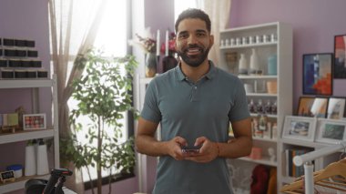 İyi dekore edilmiş bir ev dekorasyonunda cep telefonu kullanan genç adam, gülümsüyor ve çeşitli dekoratif eşyalar ve bitkilerin arasında duruyor..