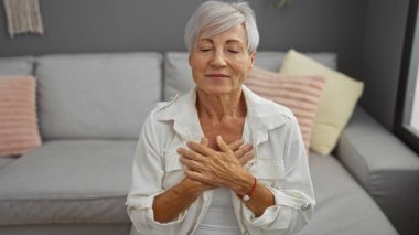 Gri saçlı olgun bir kadın, elleri göğsünde bir salonda huzur içinde, huzur ve neşe saçarak oturur.