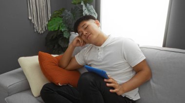 Yakışıklı genç Çinli adam, elinde bir tablet ve turuncu yastıkla şık bir oturma odasında dinleniyor.