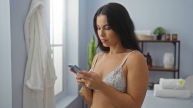 İç çamaşırlı güzel İspanyol kadın, sakin bir spa odasında akıllı telefon kullanıyor. Pencerede beyaz bir bornoz asılı.
