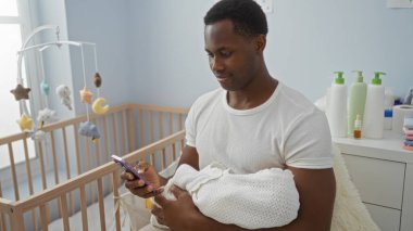 Modern yatak odasında bebeği tutan genç bir adam, akıllı telefon kullanarak, rahat bir beşiğin yanında oturmuş, huzurlu bir aile anını canlandırıyor..