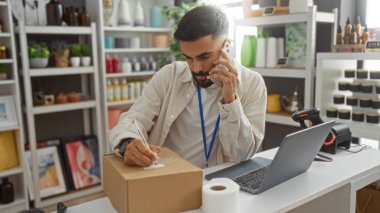 Ev dekorasyonu mağazasında sakallı genç adam, telefonla konuşuyor ve kutu etiketinde yazıyor, dizüstü bilgisayarı ve tarayıcısı var.