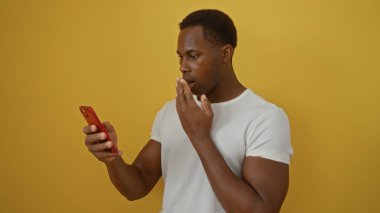 Akıllı telefon tutan genç adam sarı arka plana şaşırmış gibi görünüyor. Beyaz gömlekli, parlak duvarın üzerinde dışa dönük bir duruşu var..