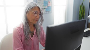 Gündüz vakti modern ofis ortamında çalışkan, yaşlı, kır saçlı bir kadın bilgisayarın başında çalışıyor..