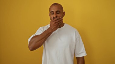 Beyaz tişörtlü, sarı arka planda esneyen yakışıklı Afro-Amerikan bir adam.