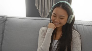 Oturma odasında kulaklıkla müzik dinleyen genç Çinli bir kadın. İçerideki rahat bir kanepede gülümsüyor ve rahatlıyor.