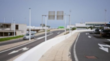 Açık gökyüzü altında arabaları olan şehir dışı bir yolun bulanık görüntüsü, arka planda binalar ve tabelalarla çözünmüş bir bokeh etkisi gösteriyor.